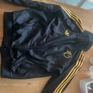 Svart träningsjacka från Adidas - Säljer en svart träningsjacka från Adidas med gula ränder längs ärmarna och en dragkedja framtill. Jackan har det klassiska Adidas-loggan på bröstet och är perfekt för en sportig look.  Tröjan är en collab med fotbollsspelaren jude Bellingham. Den är i gott skick och köptes för 1.100 