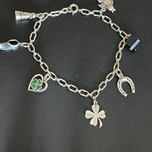 Silver charmarmband med lyckobringande berlocker - Charmigt silverarmband med flera lyckobringande berlocker, inklusive en fyrklöver, hästsko och hjärta med grön detalj. Perfekt för att ge en personlig touch till din stil.