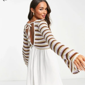 Klänning med stickad detalj - Klänning med randigt stickat mönster i brunt, vitt och lila. Den har långa ärmar och en v-ringad design med knytning i ryggen. Kjoldelen är vit bomullsmaterial.  använd 1 gång, nypris 449kr