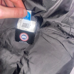 Svart dunväst från Canada Goose - Säljer en svart dunväst från Canada Goose med deras ikoniska logga på bröstet. Västen har en stilren design med knappar framtill och är perfekt för kyligare dagar. Den är tillverkad i slitstarkt material och ger bra värmeisolering.