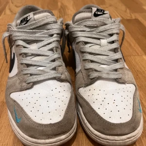 Nike sneakers i grått och vitt - Snygga Nike sneakers med en kombination av grå mocka och vitt skinn. Skorna har en svart Nike-logga på sidan och en liten blå detalj framtill. De har klassisk snörning och en bekväm passform.