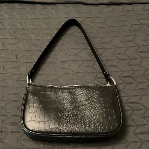 Svart axelväska från H&M Divided - Snygg svart axelväska i krokodilpräglat mönster från H&M Divided. Väskan har en dragkedja upptill och en enkel axelrem. Perfekt för att ge en stilren touch till din outfit.