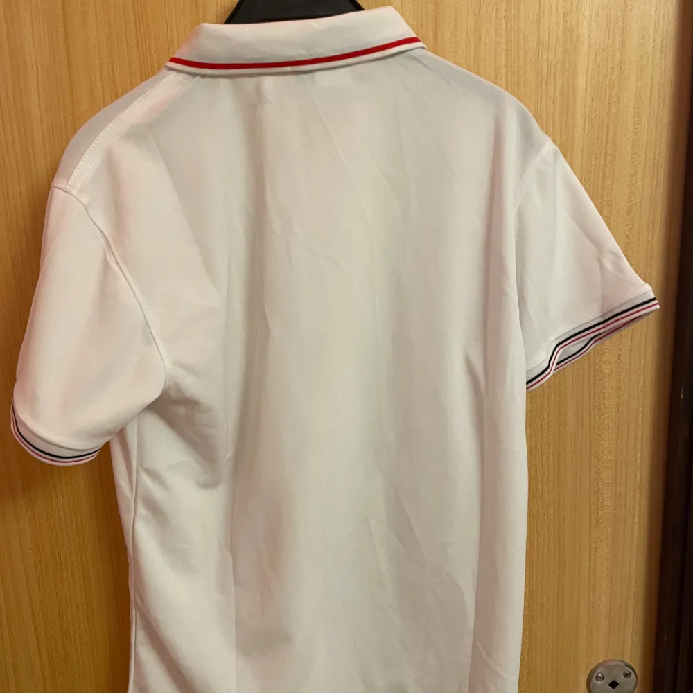 Säljer en stilren vit pikétröja från Hanyi Classic Fashion med Moncler-inspirerad logga på bröstet. Tröjan har korta ärmar och en klassisk krage med röd och svart randdetalj. Perfekt för en avslappnad men ändå snygg look.. Paidat.