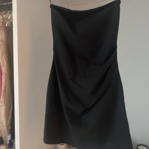 Svart axelbandslös klänning från Pull&Bear - Snygg svart axelbandslös klänning från Pull&Bear. Perfekt för en stilren look med sin korta längd och minimalistiska design. Klänningen har en dragkedja bak 