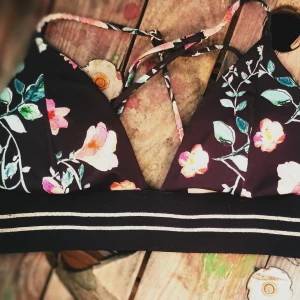 Blommig bikiniöverdel med knytband - Snygg bikiniöverdel med blommigt mönster på svart bakgrund. Den har smala knytband och en bred randig kant under bysten. Perfekt för en dag på stranden eller vid poolen.