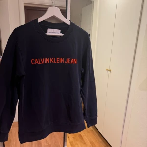 Svart sweatshirt från Calvin Klein Jeans - Säljer en stilren svart sweatshirt från Calvin Klein Jeans med orange text på bröstet. Tröjan har långa ärmar och en rund halsringning. Perfekt för en avslappnad look. Klockrent med en skjorta innanför! Skick 9/10 storlek m