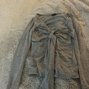 Grå långärmad topp från Gina Tricot - Säljer en grå Gina tricot topp med knut där fram, toppen har används en gång och är som ny. Köpte den precis innan hösten så jag har tyvärr inte hunnit använda den för att den är ganska tunn, och så har den blivit för liten för mig 💕