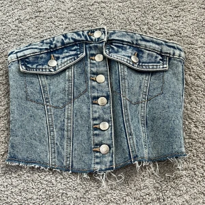 Jeans korsett från FB Sister - Snygg jeans korsett från FB Sister i en klassisk blå denim. Den har knappar framtill och fransiga kanter för en trendig look. Perfekt för att ge en edgy touch till din outfit.