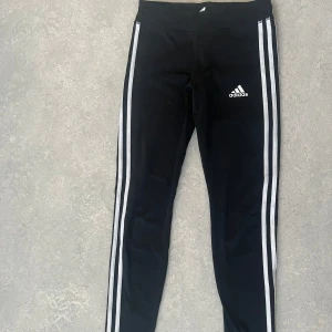 Svarta leggings från Adidas - Snygga svarta leggings från Adidas med de klassiska tre vita ränderna längs sidorna. Perfekta för träning eller en avslappnad dag. Adidas-loggan är tryckt på ena benet.