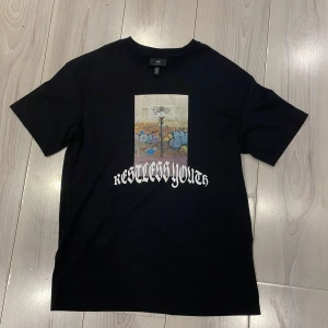 Svart t-shirt Restless Youth från H&M - Säljer en svart t-shirt från H&M med ett coolt tryck av en basketkorg och graffiti samt texten 'Restless Youth'. T-shirten har en relaxed fit och är kortärmad, Nypris 300kr. Perfekt för en avslappnad stil. 