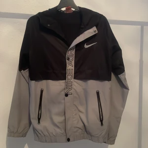 Svart och grå Nike jacka - Säljer en stilren svart och grå vindjacka från Nike med dragkedja och huva. Jackan har långa ärmar och två praktiska fickor med dragkedja. Perfekt för blåsiga dagar. Nike-loggan är broderad på bröstet.