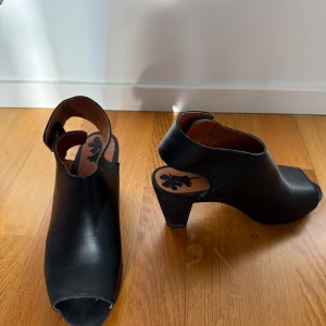 Svarta klackskor med öppen tå - Osnvända snygga svarta klackskor i skinn med öppen tå. Skorna har en praktisk knäppning baktill med tryckknappar och en stabil klack som ger extra höjd.