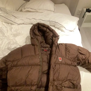Brun dunjacka från Fjällräven - Säljer en brun dunjacka från Fjällräven med huva och dragkedja. Jackan har en klassisk design med Fjällrävens logga på bröstet. Perfekt för kyliga dagar. Möts upp i stockholm