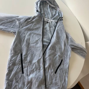 Grå vindjacka med huva - Snygg grå vindjacka från Stayhard med huva och dragkedja. Jackan har ett diskret rutigt mönster och svarta dragkedjor på fickorna. Perfekt för blåsiga dagar!