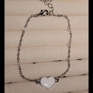 Silverfärgat armband med hjärta - Charmigt silverfärgat armband med en vit hjärtformad detalj i mitten. Armbandet har en fin kedja och ett praktiskt lås för enkel användning. Perfekt för att ge en romantisk touch till din stil.