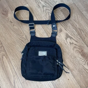 Svart axelväska från Jescinsio - Säljer en svart axelväska från Jescinsio med justerbara remmar och flera dragkedjefickor. Väskan har en metallplatta med logotyp på framsidan och är perfekt för att bära dina dagliga nödvändigheter. Enkel och stilren design som passar till många tillfällen.