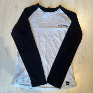 Vit och svart långärmad tröja från Dickies - Säljer en vit och svart långärmad tröja från Dickies med tryck på bröstet. Tröjan har en klassisk raglan-design med kontrasterande svarta ärmar och vit kropp. Perfekt för en avslappnad stil.
