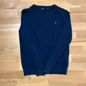 Mörkblå kabelstickad tröja från Gant - Säljer en stilren mörkblå kabelstickad tröja från Gant. Tröjan har ett broderat emblem på bröstet och är tillverkad i ett mjukt material. Perfekt för kyligare dagar.