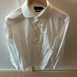 Vit skjorta från Riley - Klassisk vit skjorta från Riley med krage och knappar framtill. Perfekt för en stilren look. Skjortan har långa ärmar och är tillverkad i ett mjukt material.