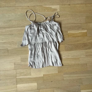 Beige axelbandslös topp - Säljer en stilren beige axelbandslös topp med smala axelband och ett lager av volang. Perfekt för varma sommardagar eller en avslappnad kväll. Toppen har en luftig och lätt känsla.