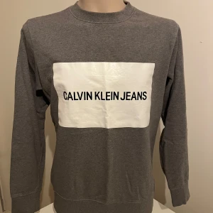 Grå sweatshirt från Calvin Klein Jeans - Säljer en stilren grå sweatshirt från Calvin Klein Jeans med en stor vit logga på framsidan. Tröjan har långa ärmar och en rund halsringning. Perfekt för en avslappnad look.