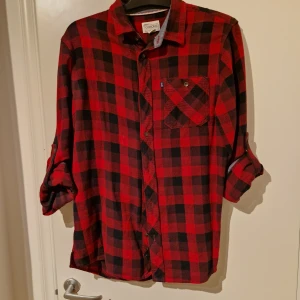 Röd och svart rutig skjorta - Snygg röd och svart rutig skjorta med klassisk krage och knappar framtill. Skjortan har en bröstficka och långa ärmar som kan rullas upp för en avslappnad look. Perfekt för en casual stil!