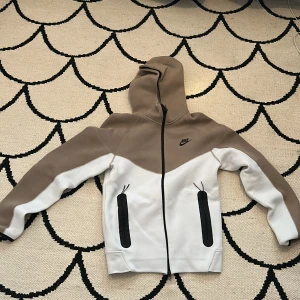 Beige och vit hoodie från Nike - Säljer en snygg beige och vit hoodie från Nike med dragkedja och huva. Jackan har två praktiska fickor med svarta dragkedjor framtill och Nike-logga på bröstet. Perfekt för en sportig look!Den har ingen fläck och är xs i vuxen storlek.