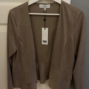 Beige jacka/kofta från Flash - Säljer en stilren beige/brun jacka från Flash med dragkedja framtill. Jackan har långa ärmar och en unik design med dubbla volanger nedtill. Perfekt för en chic look! Ganska tunn. Nypris: 599kr (köpt för 300)