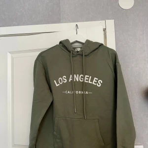 Olivgrön hoodie från H&M - Säljer en olivgrön hoodie från H&M med texten 'Los Angeles California' tryckt på framsidan. Tröjan har en klassisk känguruficka och justerbar huva med dragsko. Perfekt för en avslappnad stil.