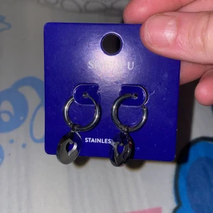 Silvriga  örhängen med hjärtdetalj från Simply U - Snygga silvriga  örhängen i rostfritt stål från Simply U. De har en rund form med en charmig hjärtdetalj som hänger ned. Perfekta för att ge en stilren touch till din outfit.