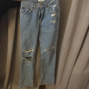 Blå jeans från Pull&Bear - Säljer ett par blå jeans från Pull&Bear med slitna detaljer. De har en klassisk femficksdesign och knappgylf. Perfekta för en avslappnad stil.