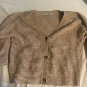 Beige stickad kofta med knappar - Säljer en mysig beige stickad kofta från 157. Koftan har en v-ringad design och tre stora knappar framtill. Perfekt för kyliga dagar och lätt att matcha med olika outfits. Passar bra för både vardag och avslappnade tillfällen.