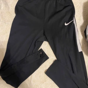 Svarta träningsbyxor från Nike - Säljer ett par svarta träningsbyxor från Nike med elastisk midja och vit logga på sidan. Byxorna har en smal passform och är perfekta för sport och träning. De har även dragkedjor vid bensluten för extra komfort, så dem är väldigt lätt och sköna att ha på. Barn storlek, 147-158 för barn mellan 12-13 år gamla. 