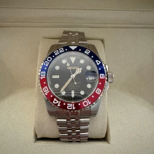 Seiko GMT Pepsi - Säljer en Seiko klocka med en iögonfallande blå och röd bezel. Klockan har en svart urtavla med tydliga markeringar och datumvisning. Armbandet är i silverfärgat stål med en klassisk länkdesign. Perfekt för den som vill ha en stilren och funktionell klocka.