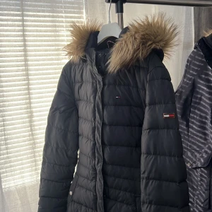 Svart dunjacka med pälskrage från Tommy Hilfiger - Snygg svart dunjacka från Tommy Hilfiger med en  pälskrage. Jackan har en lång passform och är perfekt för kalla dagar. Den har en dragkedja framtill och långa ärmar. En stilren och varm jacka för vintern. Storlek: L, men den är liten i storleken så passar M