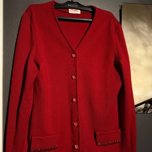 Röd kofta med stickat material - Snygg röd kofta med knappar framtill och två fickor med dekorativa detaljer. Perfekt för att lägga till en färgklick i din garderob. Koftan har en klassisk design med långa ärmar och en v-ringad hals. Materialet är  hårt och skönt stickat ! Hör av er vid frågor och intresse! Den är inte från C&A