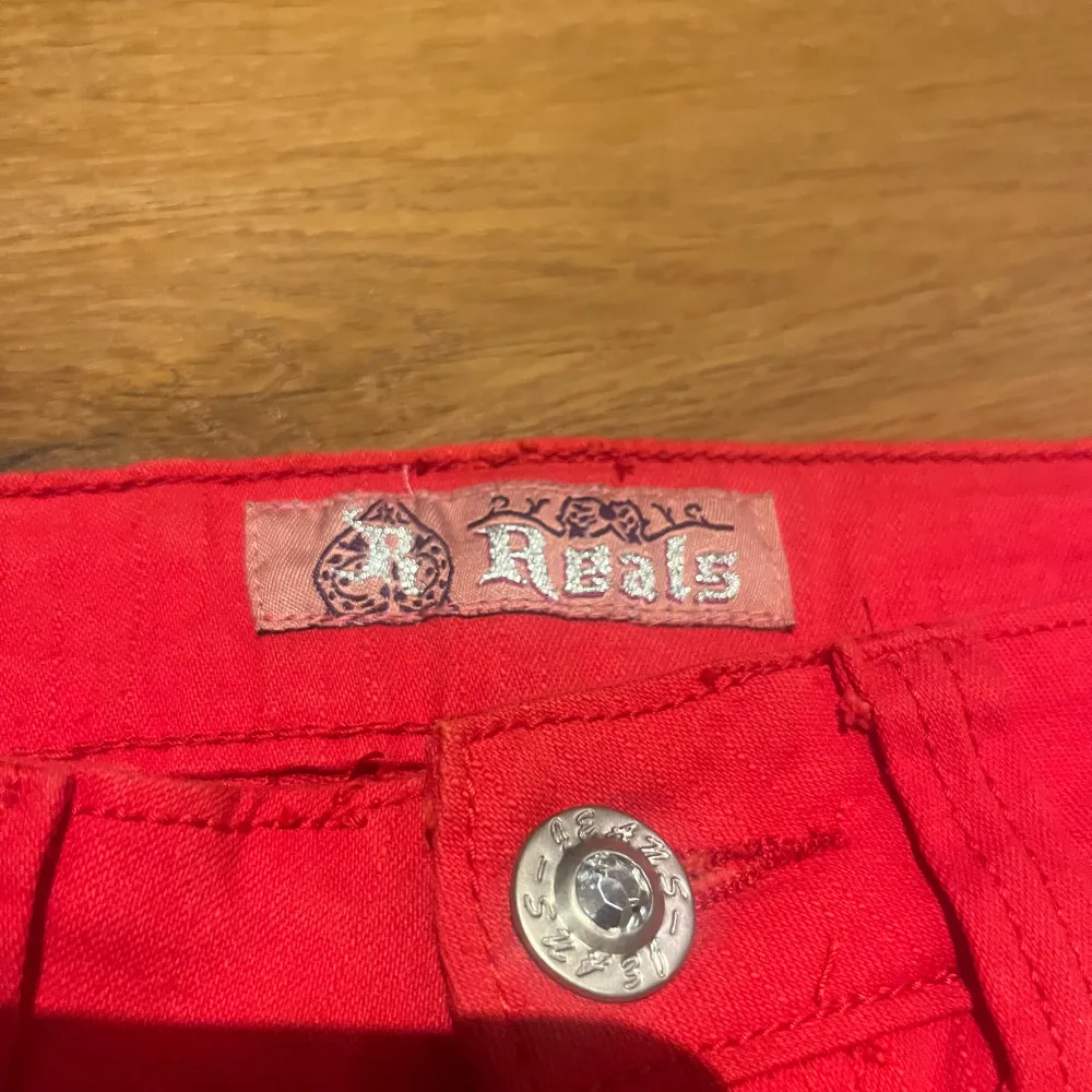 Snygga röda jeansbyxor från Reals med broderad krona på bakfickorna. Byxorna har en slim passform och är dekorerade med nitar vid fickorna. Perfekta för att sticka ut med en djärv färg och coola detaljer.. Farkut & Housut.