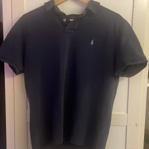 Mörkblå pikétröja från Ralph Lauren - Säljer en stilren mörkblå pikétröja från Ralph Lauren med klassisk krage och korta ärmar. Tröjan har en liten broderad logga på bröstet. Perfekt för en avslappnad men ändå snygg look. Har tappat lite färg!