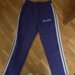 Lila träningsbyxor från Palm Angels - Snygga lila träningsbyxor från Palm Angels med vita ränder längs sidorna. Byxorna har en elastisk midja och en avslappnad passform. Säljer av hela garderoben då kläderna inte används längre.