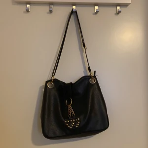 Svart axelväska med hjärtan - Snygg svart axelväska med guldfärgade detaljer och två hjärtformade hängen med nitar. Väskan har en justerbar axelrem och stora metallringar för en cool look. 