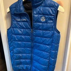 Blå quiltad väst från north sails - Snygg blå quiltad väst från North Sails med dragkedja framtill och logga på bröstet. Perfekt för kyligare dagar med sin stilrena design och bekväma passform. Insidan har en kontrasterande röd färg som ger en extra touch.Skick 9/10 bara skriva vid funderingar 