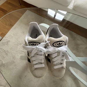 Adidas sneakers med svarta ränder - Säljer ett par klassiska Adidas campus sneakers i beige/grå med svarta ränder. Skorna har en gummisula och snörning framtill. Perfekta för en avslappnad stil. Använda ett par ggr. 