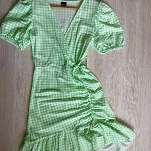 Grön rutig omlottklänning från Gina Tricot - Säljer en söt grön rutig omlottklänning från Gina Tricot. Klänningen har korta puffärmar och en volangdetalj vid fållen. Den knyts i midjan för en smickrande passform. Perfekt för sommarens alla tillfällen! 🌿