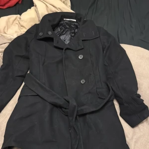 Tiger! Svart kappa med bälte - Snygg svart kappa från tiger of Sweden med knappar och bälte i midjan. Perfekt för kyligare dagar med sin stilrena design och långa ärmar. Fodrad insida för extra komfort.