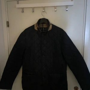 Svart quiltad jacka från Barbour - Säljer en stilren svart quiltad jacka från Barbour med klassiskt rutigt foder. Jackan har långa ärmar och stängs med knappar. Perfekt för höst och vår med sin tidlösa design och bekväma passform.