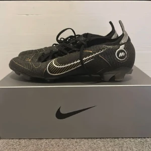 Nike Mercurial Vapor 16 Elite  - Snygga svarta fotbollsskor från Nike med vita och guldiga detaljer. Skorna har snörning och är designade för optimal hastighet på planen. Perfekta för fotbollsspelare som vill ha stil och funktionalitet.