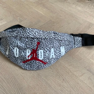 Mönstrad magväska från Jordan - Säljer en cool magväska från Jordan med ett  svartvitt mönster och den ikoniska röda loggan. Väskan har en justerbar rem och dragkedja för säker förvaring. Perfekt för att bära med sig det viktigaste! Lite fläck på baksidan, syns inte när mannar den men därav det låga priset. 