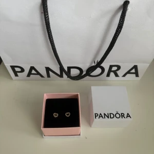Pandora örhängen + äkthetsintyg  - Säljer ett par eleganta hjärtformade örhängen från Pandora. De är i 925 silver med kristal stenar, används få gånger och i bra skick! Silverputsade inan försäljning💗