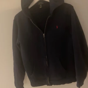  Ralph Lauren hodie  - Säljer en snygg mörkblå hoodie från Ralph Lauren med dragkedja och den klassiska röda loggan på bröstet. Perfekt för en avslappnad stil. Hoodien har långa ärmar och en bekväm passform.Storlek xl barn altså cirka M vanlig storlek 