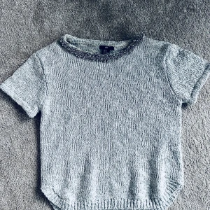 Grå stickad topp med pärlor - Snygg grå stickad topp med korta ärmar och dekorativa pärlor runt halsringningen. Perfekt för att ge en touch av elegans till din outfit. Passar bra till både jeans och kjol.